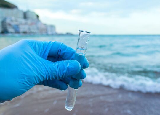 Behandschuhte Hand hält ein Reagenzglas mit klarem Wasser; im Hintergrund Meeresufer mit flacher Welle